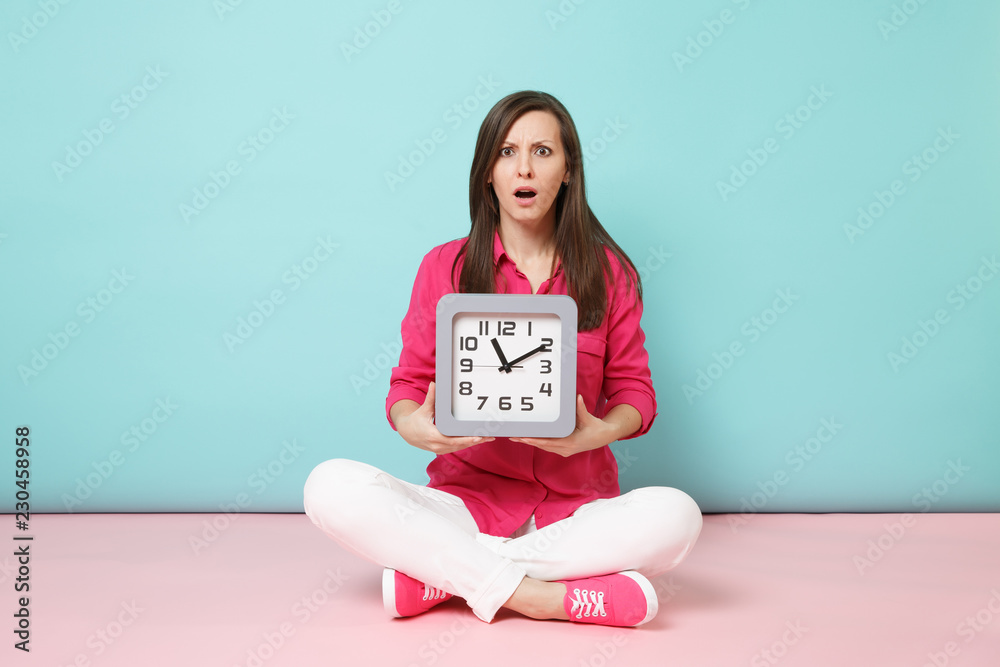 Full length portrait young woman in rose shirt blouse white pants sitting on floor hold clock isolated on bright pink blue pastel wall background studio. Fashion lifestyle concept. Mock up copy space. Full length portrait young woman in rose shirt blouse white pants sitting on floor hold clock isolated on bright pink blue pastel wall background studio. Fashion lifestyle concept. Mock up copy space.