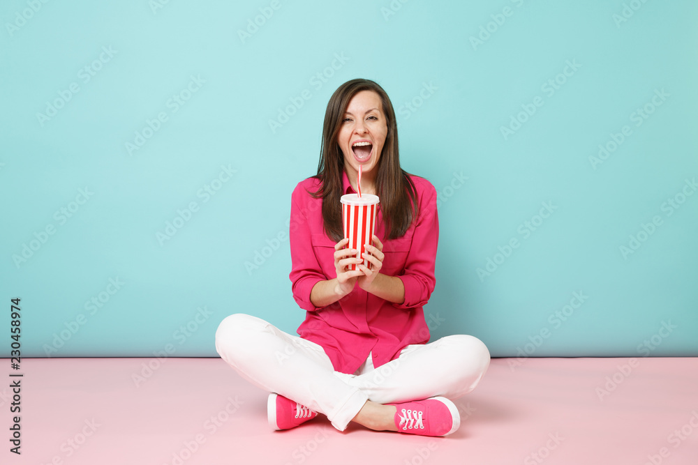 Full length portrait woman in rose shirt blouse white pants sitting on floor hold cup of soda isolated on bright pink blue pastel wall background studio. Fashion lifestyle concept. Mock up copy space. Full length portrait woman in rose shirt blouse white pants sitting on floor hold cup of soda isolated on bright pink blue pastel wall background studio. Fashion lifestyle concept. Mock up copy space.