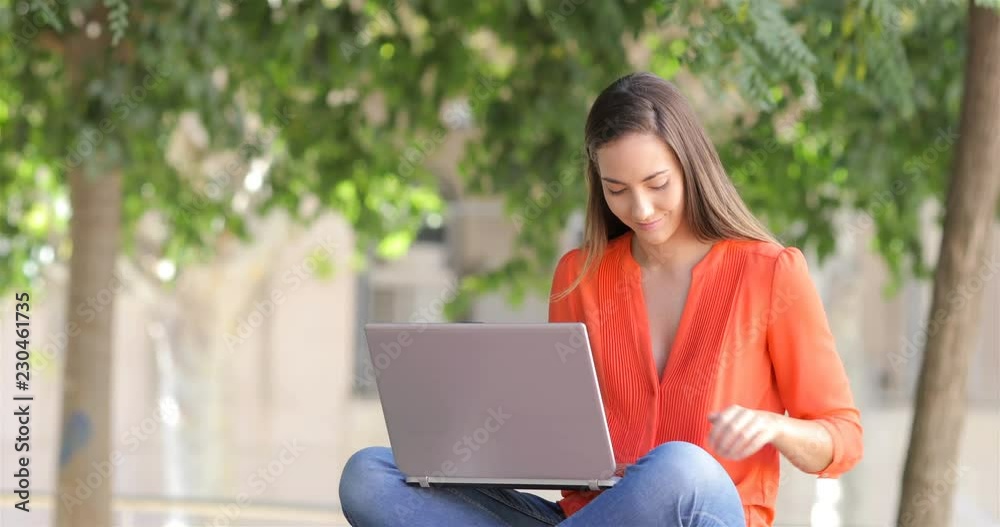 Happy woman receiving a phone message while is using a laptop sitting on a bench in a park Happy woman receiving a phone message while is using a laptop sitting on a bench in a park