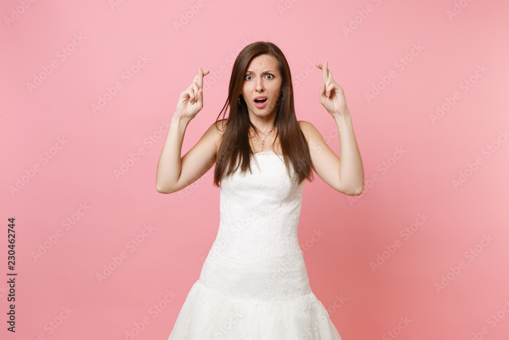 Portrait of scared bewildered bride woman in white wedding dress waiting for special moment, keeping fingers crossed isolated on pastel pink background. Wedding concept. Copy space for advertisement. Portrait of scared bewildered bride woman in white wedding dress waiting for special moment, keeping fingers crossed isolated on pastel pink background. Wedding concept. Copy space for advertisement.
