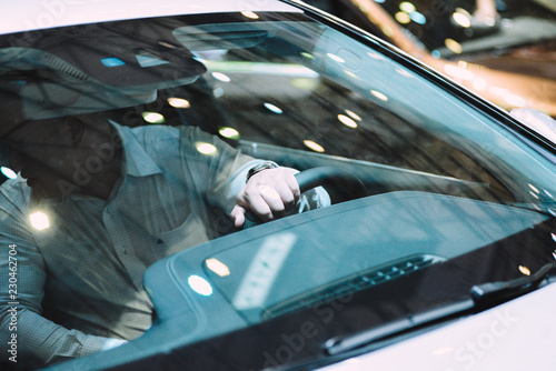 Viewed from the outside of unidentified man sitting in a car, his left hand on the steering wheel. Windshield has more bokeh light