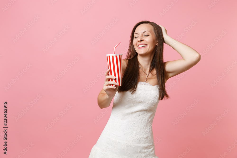 Portrait of smiling bride woman with closed eyes in white wedding dress keeping hand on head holding plastic cup with cola or soda isolated on pink pastel background. Wedding celebration. Copy space. Portrait of smiling bride woman with closed eyes in white wedding dress keeping hand on head holding plastic cup with cola or soda isolated on pink pastel background. Wedding celebration. Copy space.