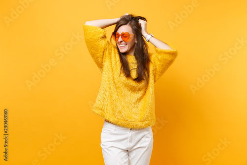 Pretty smiling young woman with closed eyes in fur sweater and heart orange glasses clinging to head isolated on bright yellow background. People sincere emotions, lifestyle concept. Advertising area.