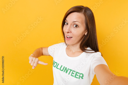 Selfie image of happy smiling satisfied woman in white t-shirt with written inscription green title volunteer isolated on yellow background. Voluntary free assistance help  charity grace work concept.