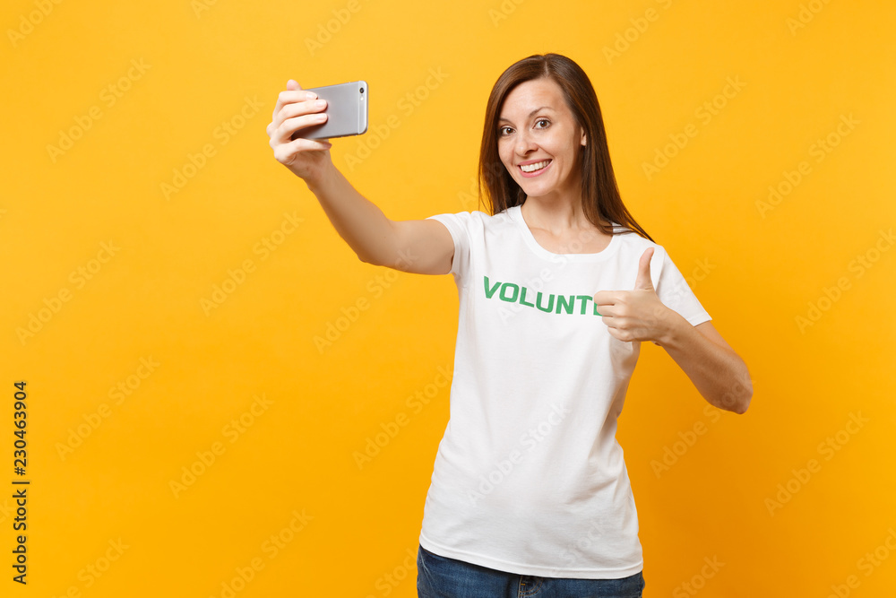 Portrait of woman in white t-shirt written inscription green title volunteer taking selfie shot on mobile phone isolated on yellow background. Voluntary free assistance help charity grace work concept