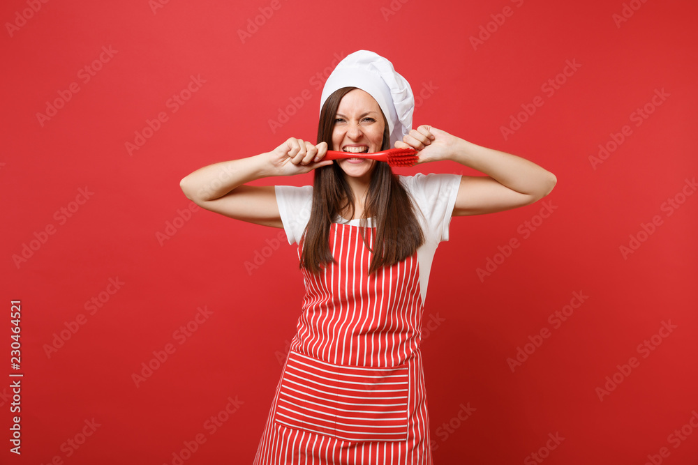 Fototapeta premium Housewife female chef cook or baker in striped apron, white t-shirt, toque chefs hat isolated on red wall background. Fun crazy woman hold in mouth brush for washing dishes. Mock up copy space concept