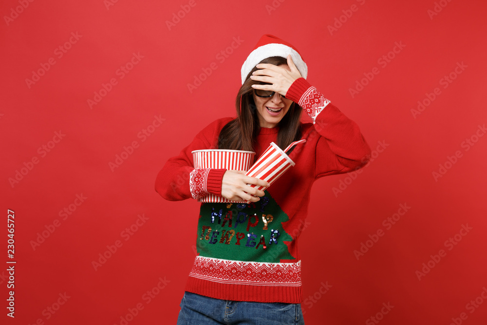 Scared young Santa girl in 3d imax glasses watching movie film hold popcorn, cup of soda covering face with hand isolated on red wall background. Happy New Year 2019 celebration holiday party concept. Scared young Santa girl in 3d imax glasses watching movie film hold popcorn, cup of soda covering face with hand isolated on red wall background. Happy New Year 2019 celebration holiday party concept.