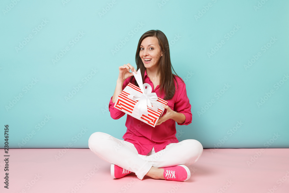 Full length portrait woman in rose shirt blouse white pants sitting on floor hold present box isolated on bright pink blue pastel wall background studio. Fashion lifestyle concept. Mock up copy space. Full length portrait woman in rose shirt blouse white pants sitting on floor hold present box isolated on bright pink blue pastel wall background studio. Fashion lifestyle concept. Mock up copy space.
