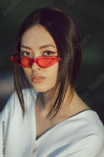 Asian woman fashion close-up portrait. Beautiful mixed race asian caucasian young girl perfect skin creartive art make-up wearing red sunglasses trendy white japanese kimono standing outdoors in © Buyanskyy Production