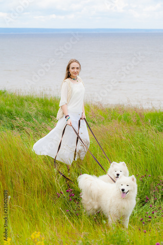 young beautiful woman standing with two big white shaggy dogs on the background of the river in the green grass