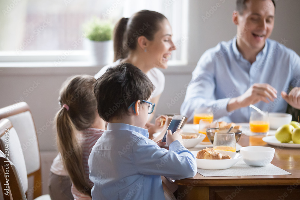 While whole family eating talking having breakfast, little preschool son in glasses holding using smartphone looking at screen. Bad habit overuse of devices and gadgets, mobile phone addiction concept While whole family eating talking having breakfast, little preschool son in glasses holding using smartphone looking at screen. Bad habit overuse of devices and gadgets, mobile phone addiction concept