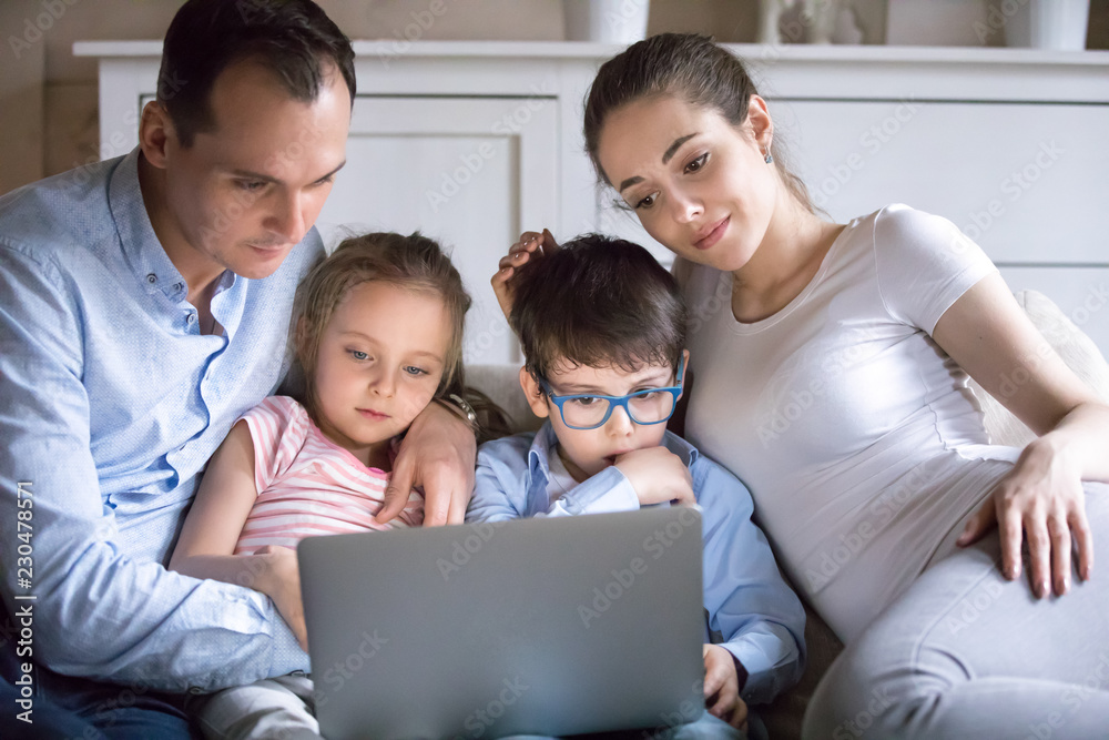 Happy family mother father embrace preschool little daughter and son relax rest in living room sitting together on sofa at home use computer watch recreational programs movie feels good and satisfied.