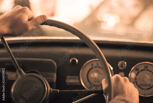 driving a vintage car; only the driver’s hand on the steering wheel are visible, the dashboard is blurred; stylized as an old sepia photo with dust and noise