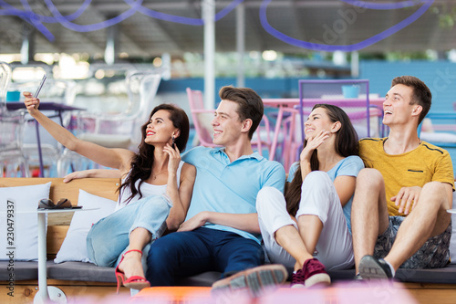 A company of good-looking friends is laughing and sitting on the bench with cushions and making selfie in the nice summer cafe. Entertainment, having good time. Friendship.