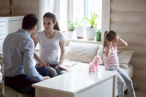 Married couple and little daughter at home wife husband sorting out their relationships arguing quarreling, small kid closed ears with hands not to hear them. Break up and divorce, kid suffers concept