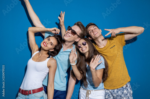 Four good-looking friends are laughing while standing in front of the blue wall having confident and happy looks. Entertainment, having good time. Friendship, relationship. © Leika production