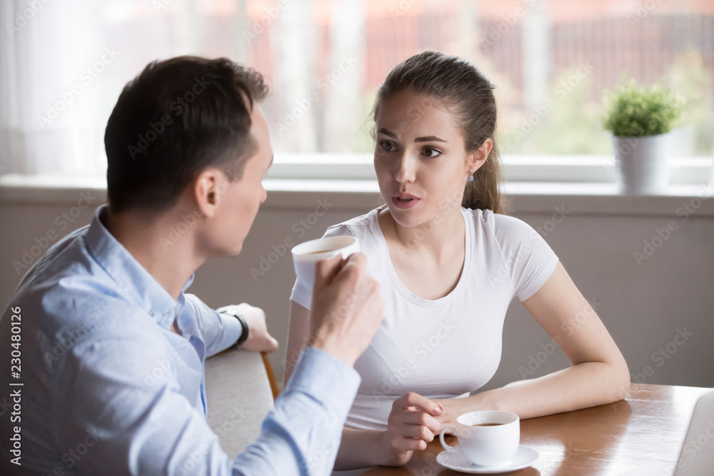 Attractive serious millennial couple in love sitting in kitchen drinking morning coffee at home. Young childfree wife and husband talking about upcoming work day or discussing about plans on weekend Attractive serious millennial couple in love sitting in kitchen drinking morning coffee at home. Young childfree wife and husband talking about upcoming work day or discussing about plans on weekend