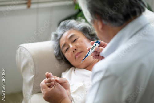 Sick Asian Senior Woman laying on couch while her husband holding and looking to temperature.