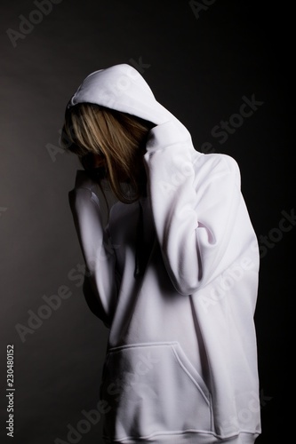 A beautiful young woman in a large white hooded sweatshirt hides her face. Studio portrait on black background