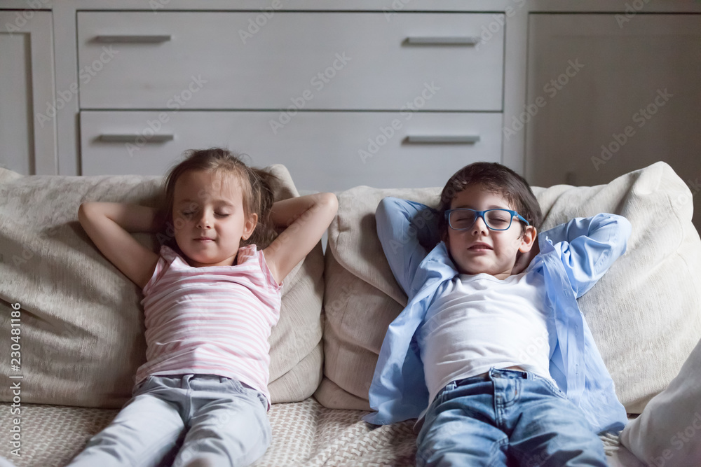 Silent preschool sister and brother sitting on sofa in living room at home. Siblings closing eyes have break breath fresh air putting hands behind heads relaxing thinking. Weekend daydreaming concept