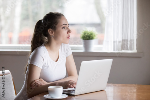 Pensive beautiful millennial woman writer sitting in kitchen at table thinking contemplating about new article for online journal. Thoughtful female using computer spend free time on weekend at home