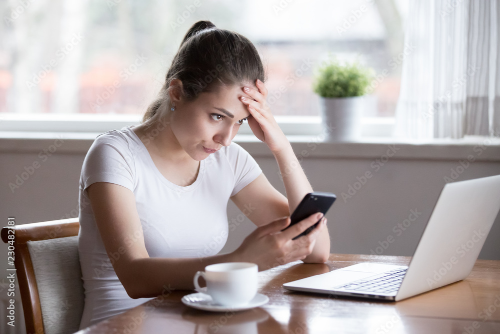 Annoyed millennial woman sitting alone in kitchen at table at home holding smartphone gadget looking at computer screen reading message notification feels irritated and upset have problems with device Annoyed millennial woman sitting alone in kitchen at table at home holding smartphone gadget looking at computer screen reading message notification feels irritated and upset have problems with device