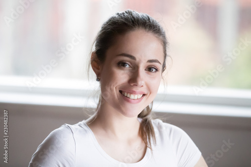Head shot of happy single millennial candid woman smiling and looking at camera at home. Portrait of beautiful female brunette girl with wide toothy smile feeling happy healthy posing alone indoors