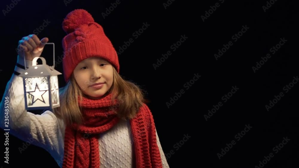 Little girl wearing knitted winter hat and scarf standing with lantern smiling at camera, over black background