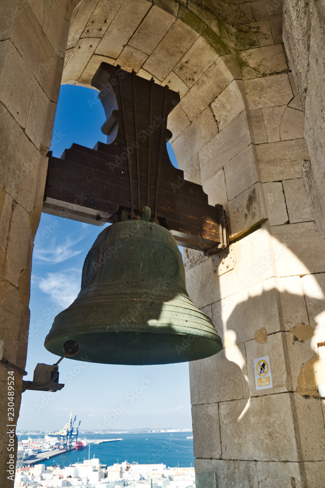 A church bell from the cathedral of Cadiz in the center of the frame suspended in its arched opening.