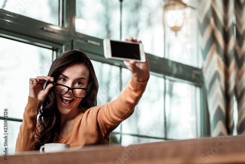 Playful mood. Delighted brunette woman looking at her gadget while taking picture