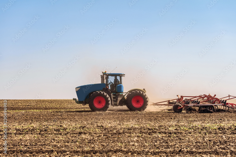 Fototapeta premium Agricultural tractor is working in the field under blue sky at daylight. Soil cultivation. Agrotechnical event that provides crumbling, loosening and partial mixing of the soil.