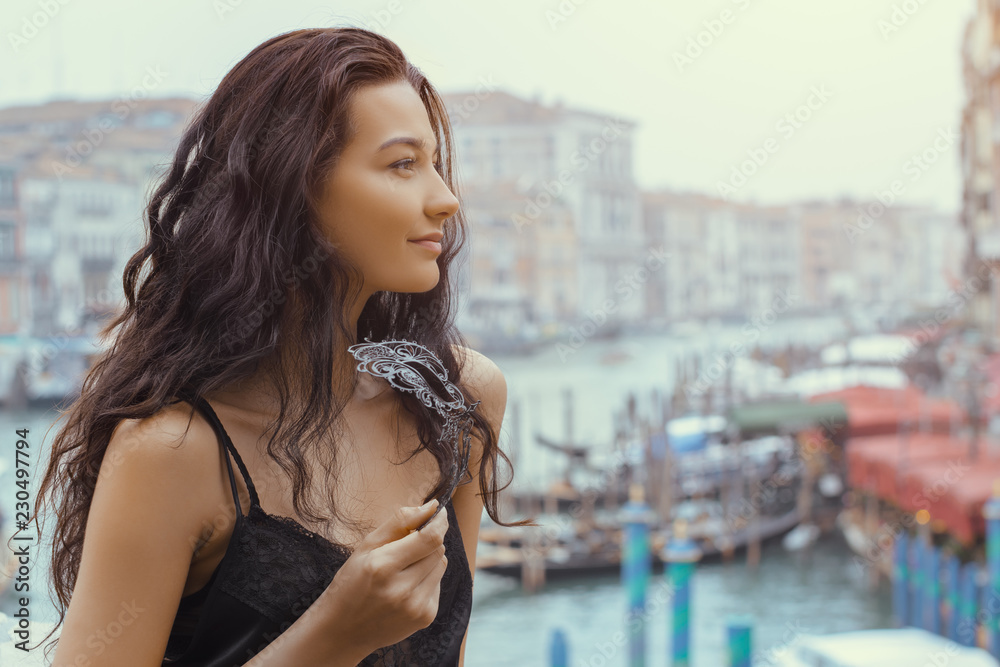 Woman with carnival mask in Venice. Attractive young sensual romantic woman standing on the pier against beautiful view on venetian chanal with boats and gondolas in Venice, Italy.