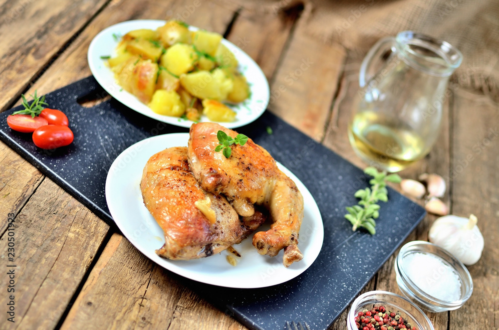 Golden grilled chicken legs on a plate and roasted potatoes, tomatoes, rosemary, oil, oregano, salt, pepper, garlic, black cutting board on a wooden table