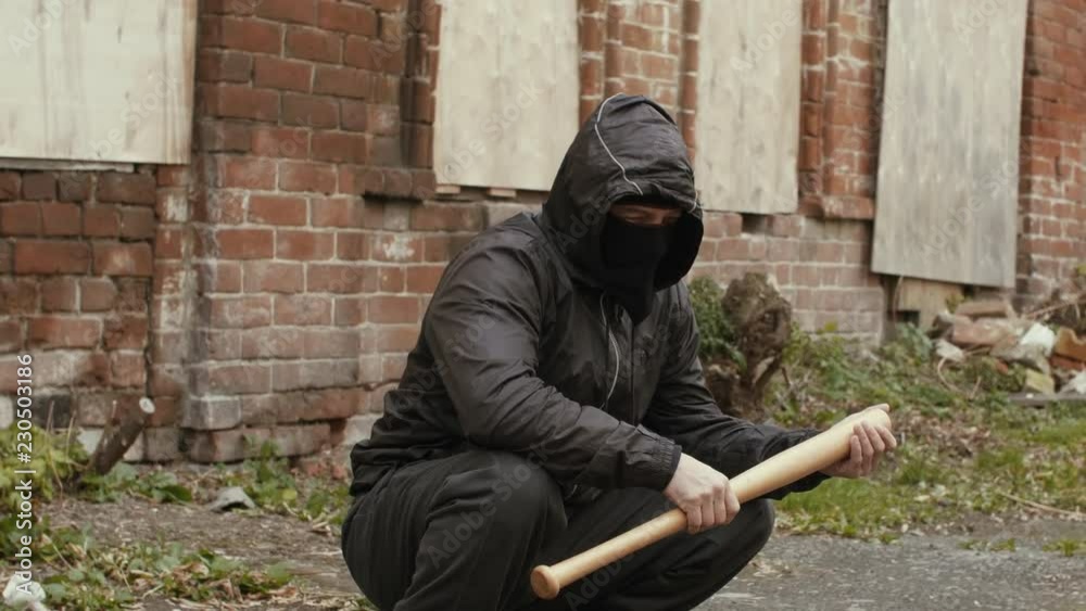 Bandit man in black mask and jacket with hood with baseball bat sits on street Bandit man in black mask and jacket with hood with baseball bat sits on street