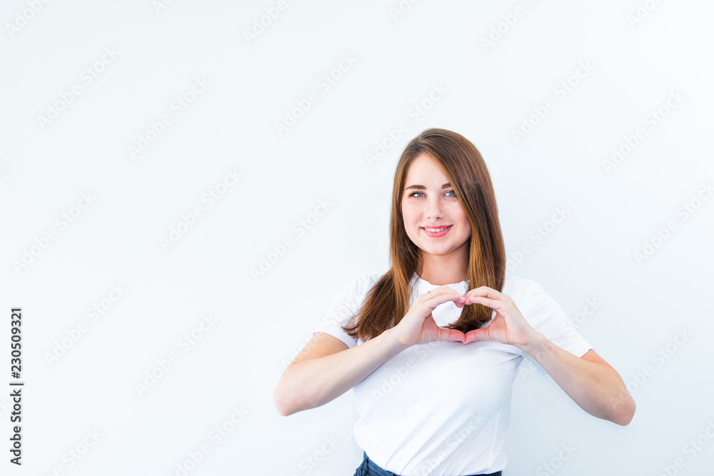Portrait of young, brunette, pretty girl in white T shirt showing with her finger heart shape, sending love. Cancer protect. Isolated on white background. Human emotions. Selective focus, copy space. Portrait of young, brunette, pretty girl in white T shirt showing with her finger heart shape, sending love. Cancer protect. Isolated on white background. Human emotions. Selective focus, copy space.