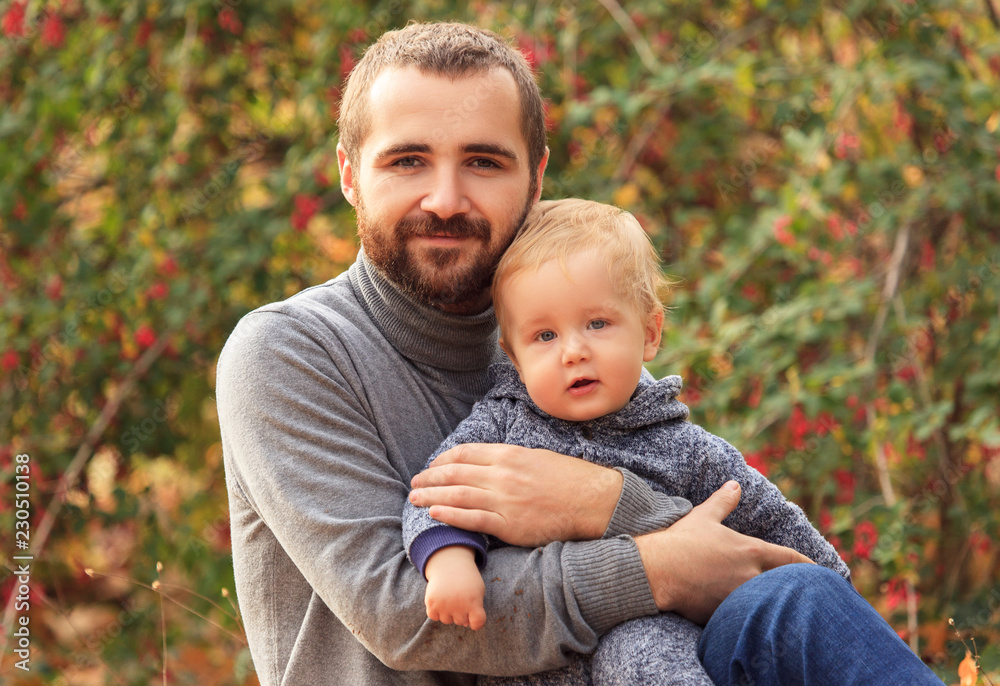 Family picnic in the autumn in the forest. Close-up portrait of a young dad with a young son among yellow foliage on the background of a green-yellow bush with berries Family picnic in the autumn in the forest. Close-up portrait of a young dad with a young son among yellow foliage on the background of a green-yellow bush with berries