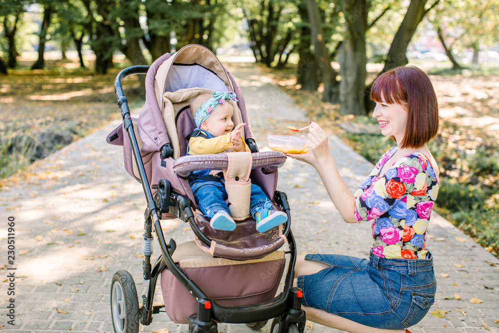 Obraz premium Young attractive mother feeding her cute baby girl, giving her first solid food, healthy vegetable pure from carrot with a plastic spoon sitting on baby stroller carriage and posing smiling