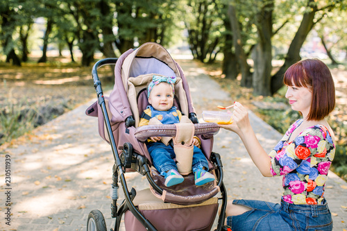 Young attractive mother feeding her cute baby girl, giving her first solid food, healthy vegetable pure from carrot with a plastic spoon sitting on baby stroller carriage and posing smiling