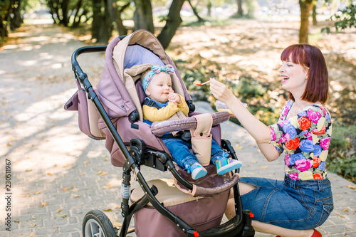 Young attractive mother feeding her cute baby girl, giving her first solid food, healthy vegetable pure from carrot with a plastic spoon sitting on baby stroller carriage and posing smiling