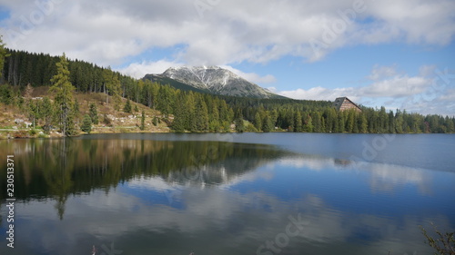 Strbske pleso and Tatra peaks visible from the back. A village located in the valley  from which tourists are moving to the Tatras. Colorful waters of a mountain pond  blue sky and unending peaks.