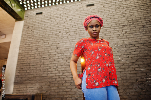 Stylish african woman in red shirt and hat posed against brick wall.