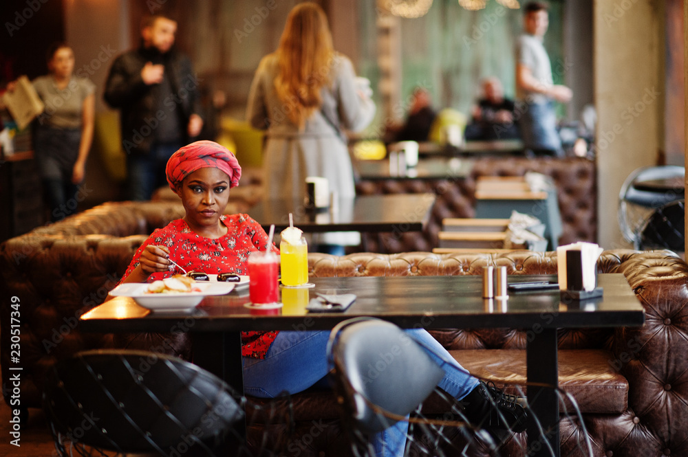 Stylish african woman in red shirt and hat posed indoor cafe, eat chocolate dessert cakes.