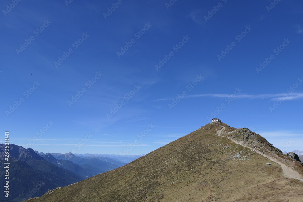 Endless expanse in the mountains. A mountain hut stands lonely on a peak in the mountains. A hiking trail leads to the hut. Blue, cloudless sky in the summer without vegetation only grass and stones