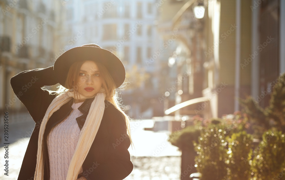 Outdoor portrait of awesome blonde girl wearing hat, scarf and coat, posing at the old street in sun glare. Empty space Outdoor portrait of awesome blonde girl wearing hat, scarf and coat, posing at the old street in sun glare. Empty space
