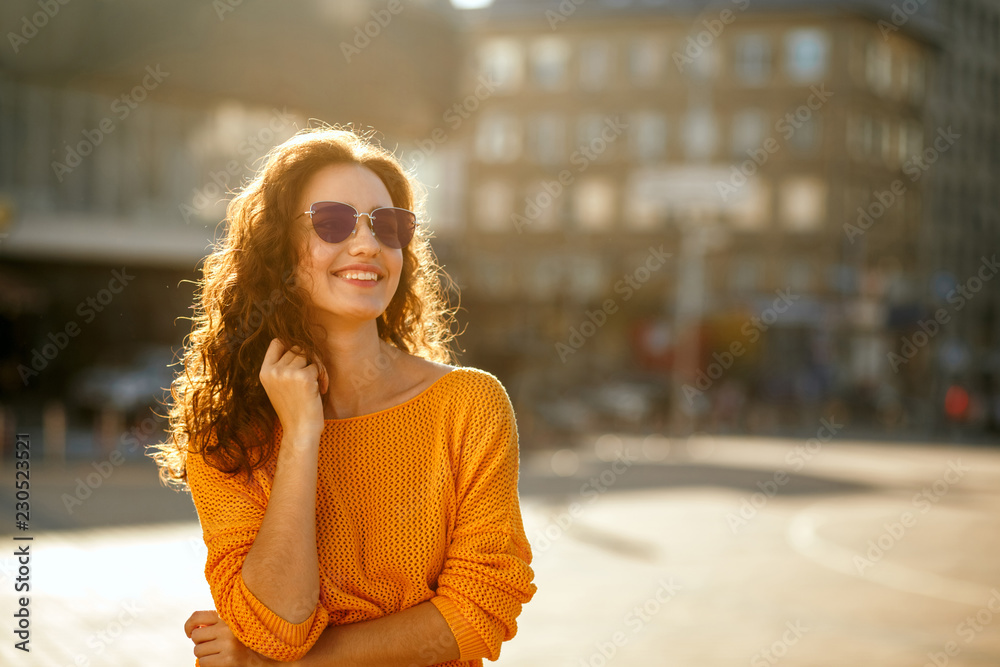 Fototapeta premium Cheerful young model with curly hair in sunglasses wearing trendy outfit and walking at the sunny street. Space for text