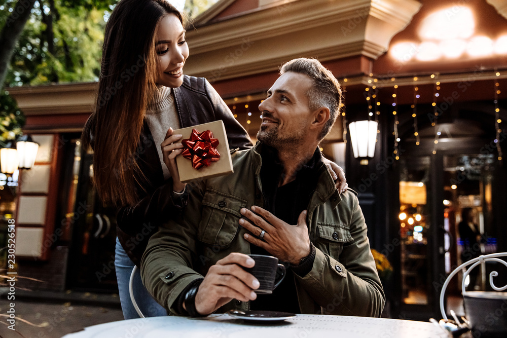 Couple. Holiday. Cafe. Woman is giving a gift box to her man. Both are in warm casual clothes smiling while sitting in the cafe outdoors