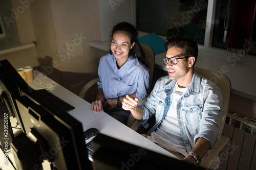 High angle portrait of two cheerful colleagues discussing work while using computer in dark office late at night, copy space