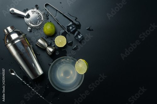 Margarita cocktail glass and bar equipments, stainless steel cocktail shaker and jigger, bar spoon with strainer, the lemons and ice tongs with ice cubes on the table photo