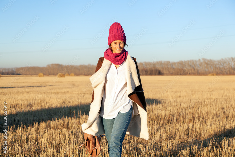The concept of livestyle  outdoor in autumn. Close up of a young woman student in a warm autumn clothes, pink knitted hat looking funny, smilling, posing for the camera, walking on field, on  forest