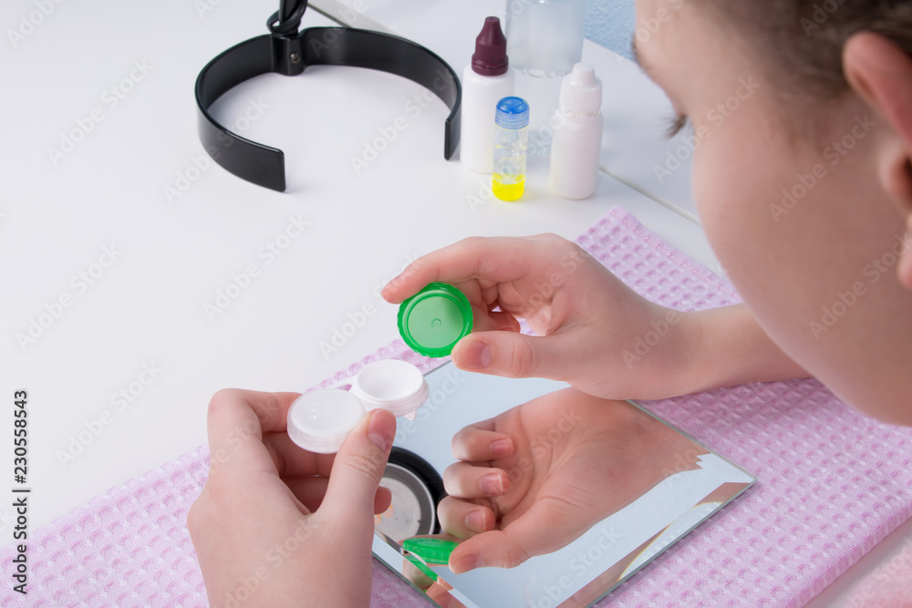 the girl, sitting at the table, opened containers for storing contact lenses, in front of a mirror the girl, sitting at the table, opened containers for storing contact lenses, in front of a mirror
